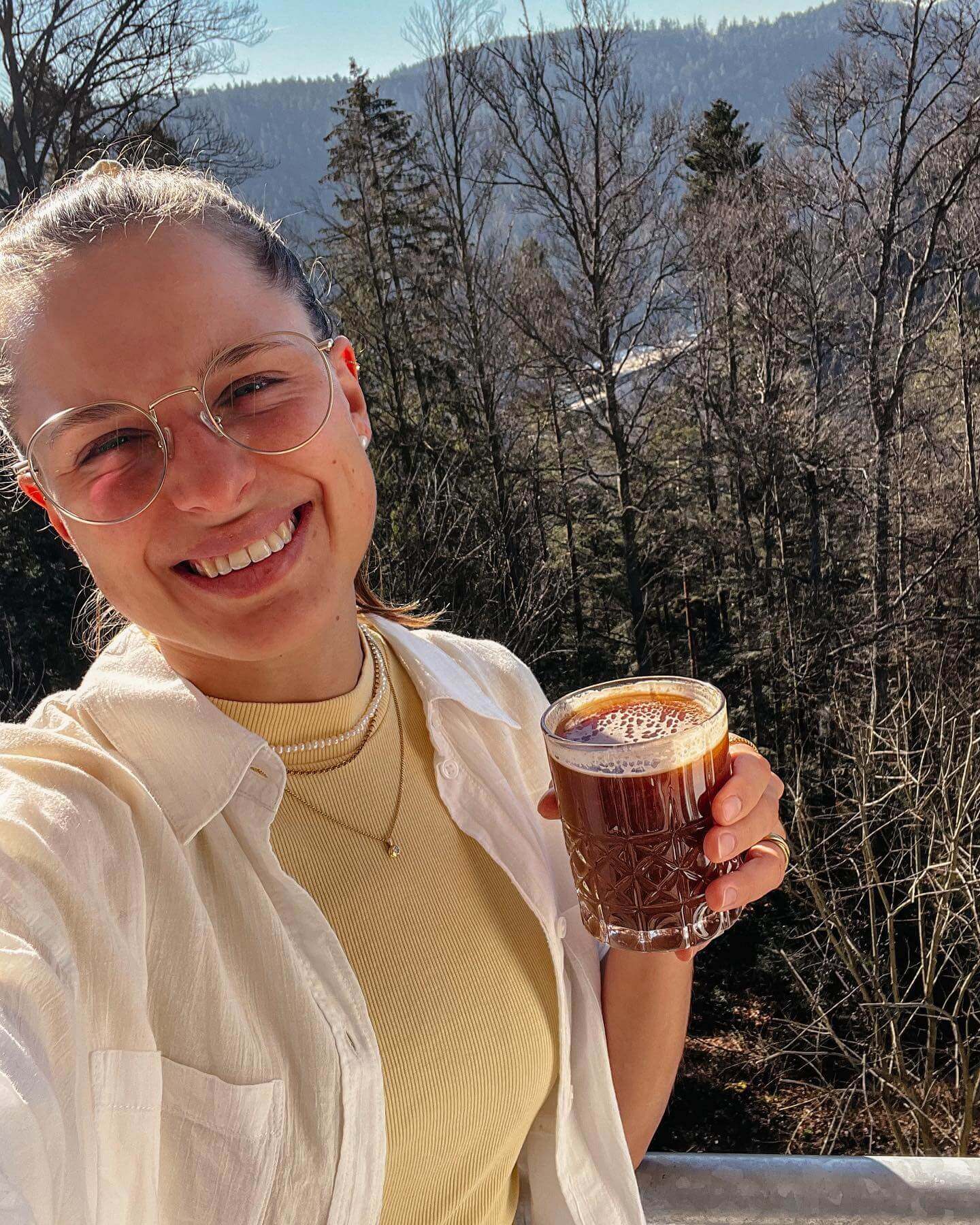 Eine Frau mit Brille und zurückgebundenen Haaren lächelt. Sie trägt ein gelbes Oberteil und eine weiße Bluse. In ihrer Hand hält sie ein Glas mit hajoona Balance. Hinter ihr sind Bäume und Berge zu sehen.