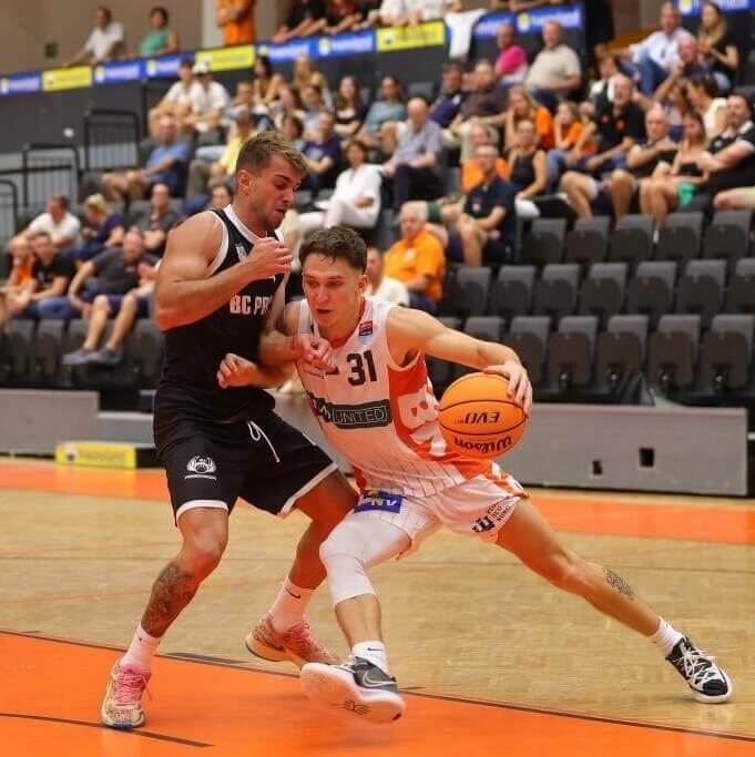 Zwei Basketballspieler im Duell. Der Profi-Basketballer Lukas Reichle trägt ein weißesn Trikot mit orangen Streifen, er dribbelt einen orangefarbenen Ball. Der andere Spieler im schwarzen Trikot versuch ihn zu stoppen. Der Boden ist orange, im Hintergrund sitzen Zuschauer.